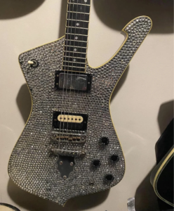 Close-up of a guitar body showing rhinestones attached with hot glue being carefully removed using a plastic guitar pick without damaging the glossy finish